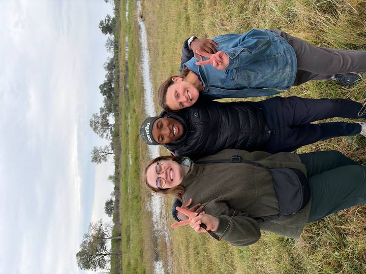 Three smiling travellers pose with peace signs against a marshy grassland and shallow water backdrop