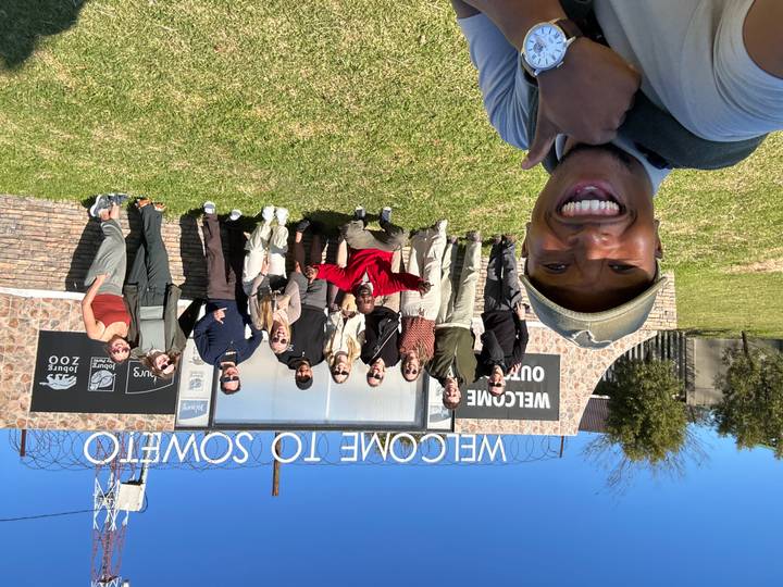 Large tour group smiles in front of a Welcome to Soweto sign on a sunny day