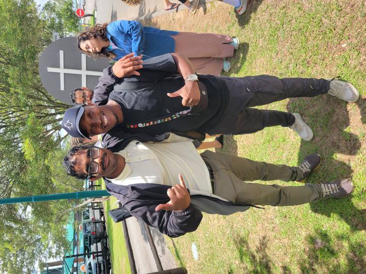 Two friends pose together outdoors during a sunny gathering, flashing smiles and thumbs-up gestures.