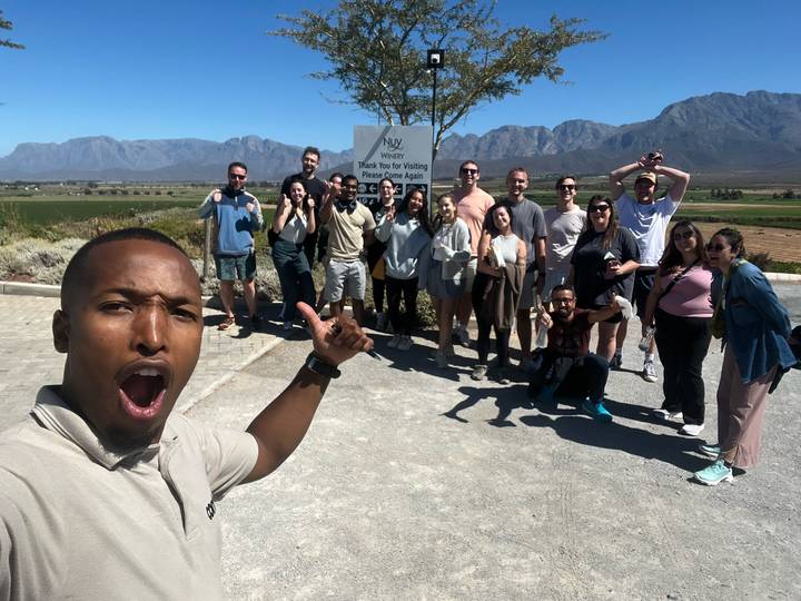 A lively group poses by a wine estate sign with vineyards and rugged mountains under a bright blue sky.
