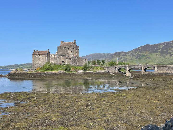 Stone Eilean Donan Castle sits on a tidal island reflected in calm waters with a small bridge.