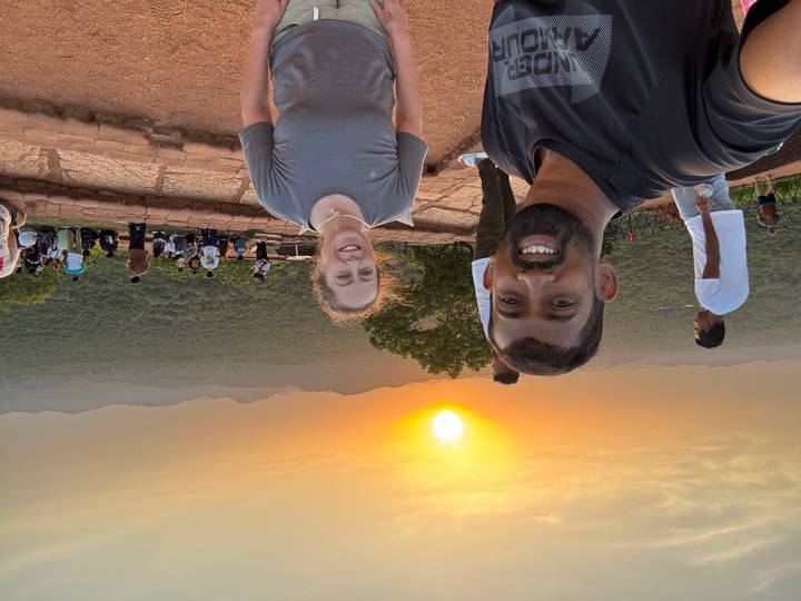 Two smiling travellers take a sunset selfie atop a stone terrace overlooking forested hills and distant mountains.