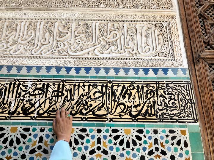Close-up of ornate Arabic calligraphy carved in stone and colorful mosaic tiles with a hand touching the wall.