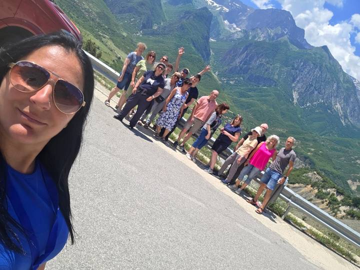 Smiling tour group poses on a mountain road with green Balkan peaks behind them.