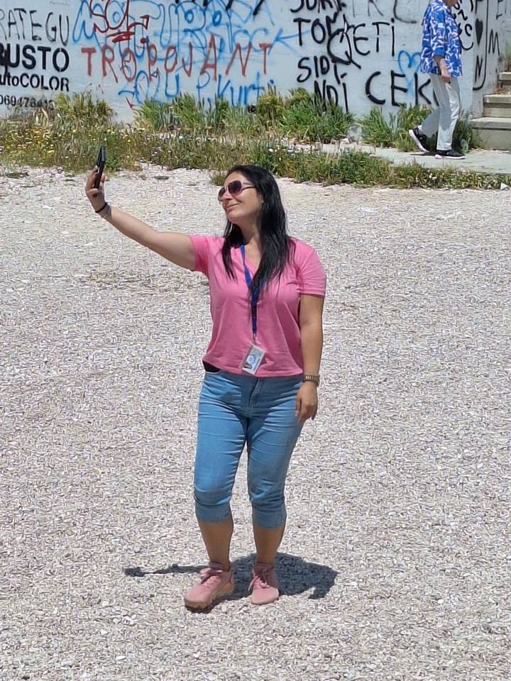 A woman in pink top takes a selfie against a gravel surface under bright sun.