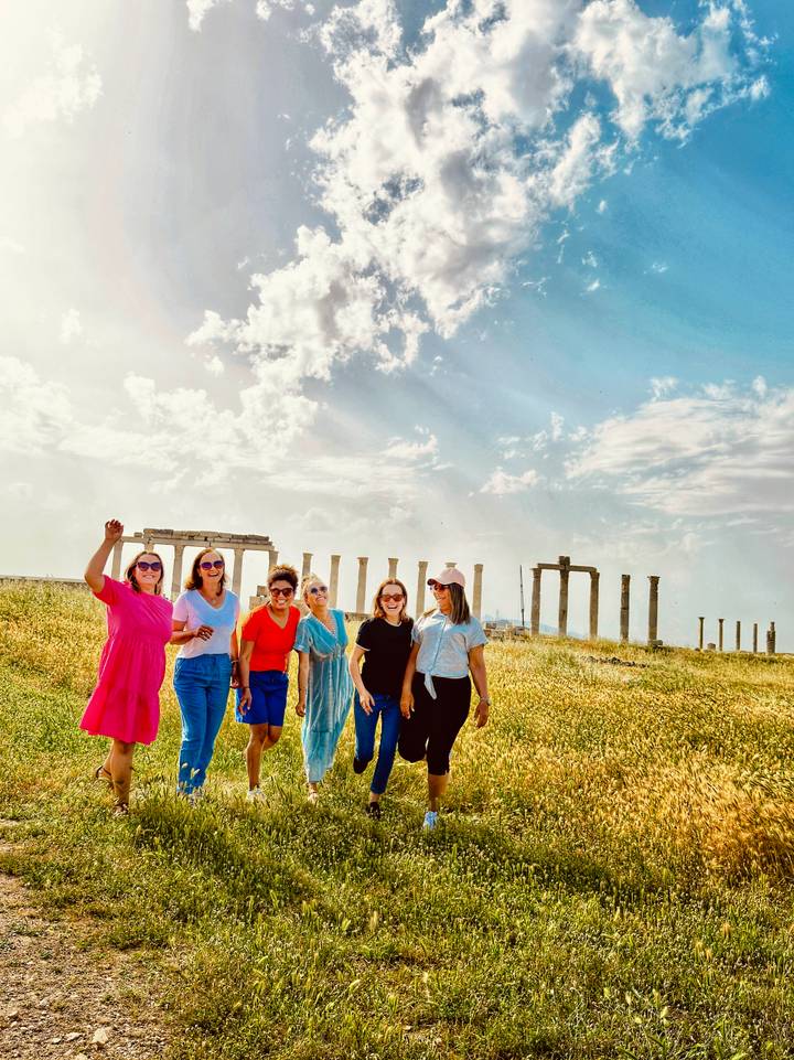 Group of friends pose cheerfully in a grassy archaeological field dotted with standing columns.