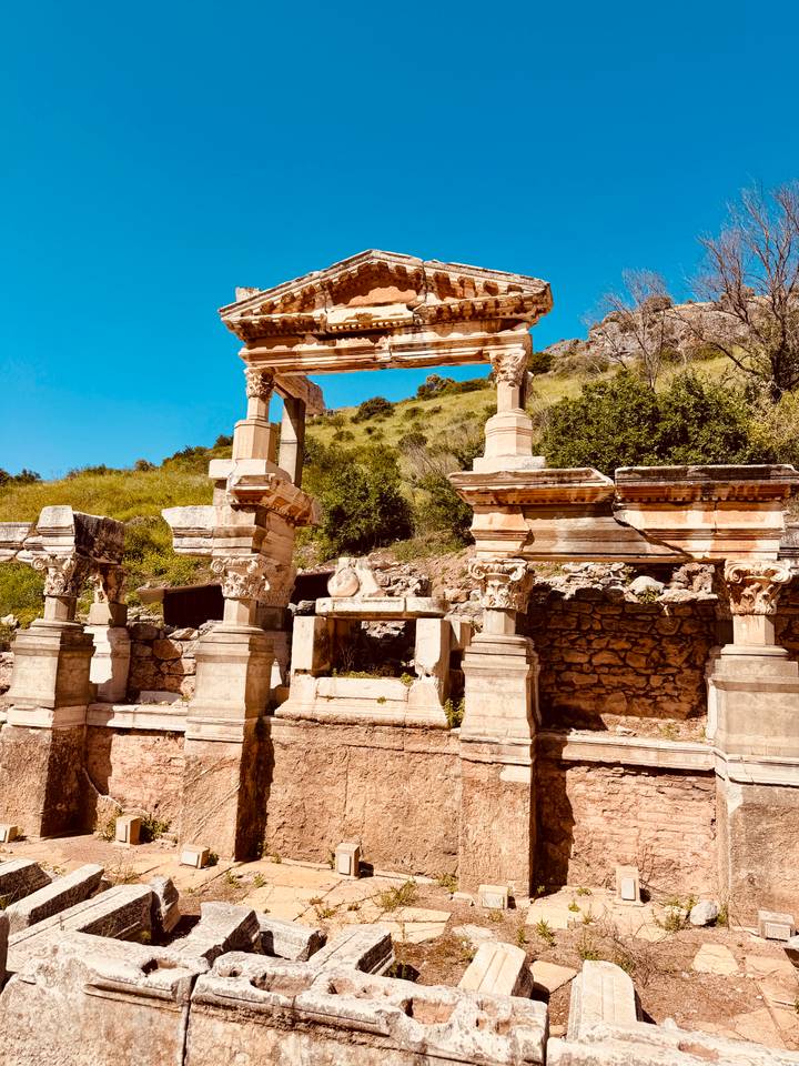Sun-bleached ruin with decorative columns set against a green hillside and vivid blue sky.