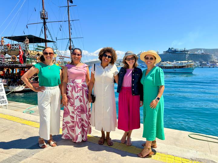 Smiling women lined up on a sunny quayside with tour boats and a cruise ship behind them.