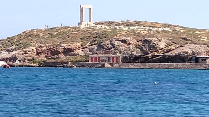 Ancient portal ruin (Portara) atop rocky hill overlooking vivid blue Aegean Sea.