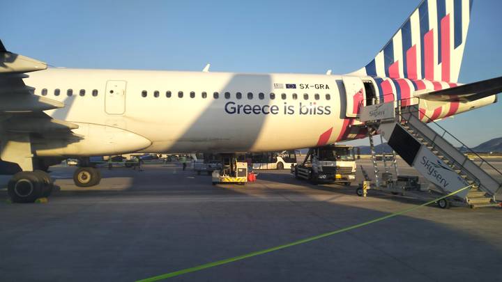 Passenger jet with colourful tail parked on tarmac displaying slogan 'Greece is bliss'.