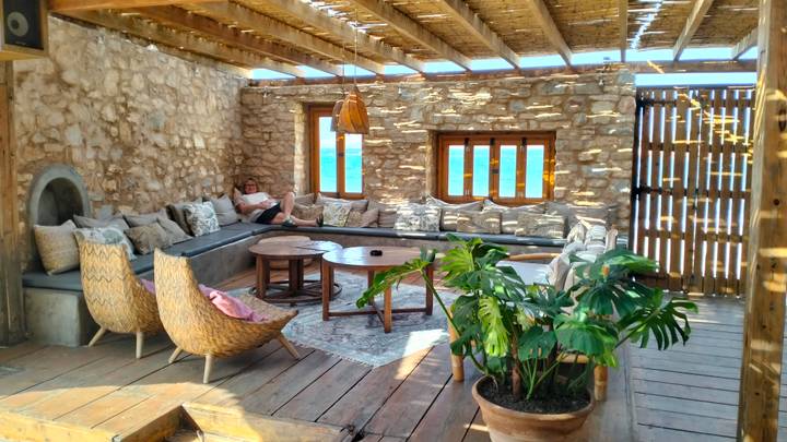 Cozy rustic lounge with sea view windows, rattan chairs and guest reading on sofa.