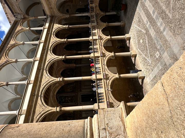 Stone cloister or palace courtyard with multiple arched levels and visitors walking along the balcony.