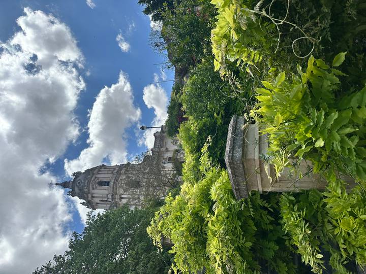 Baroque bell tower rising above lush green foliage and bright clouds in a hillside garden.