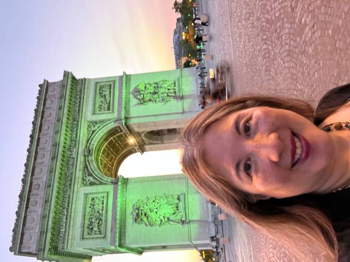 Woman taking an evening selfie with the illuminated Arc de Triomphe glowing green behind her.