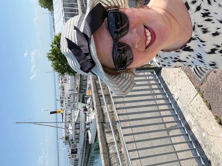 Woman in sunhat and sunglasses taking a close-up selfie with a marina full of docked boats and blue water in the background.