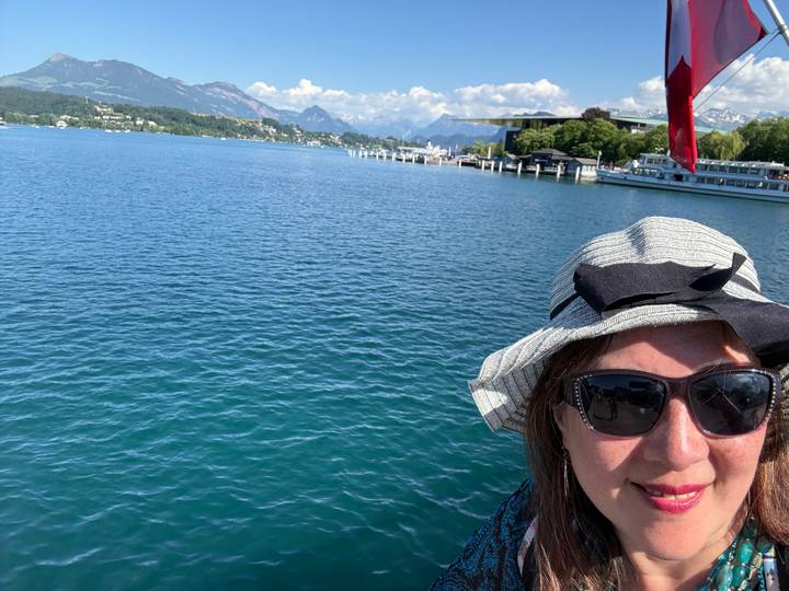 Partial selfie of a woman on a boat with deep blue lake water and snow-tipped mountains in the distance.
