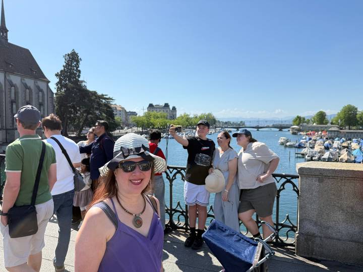 Tourists standing on a bridge overlooking a river marina filled with small boats on a bright sunny day.