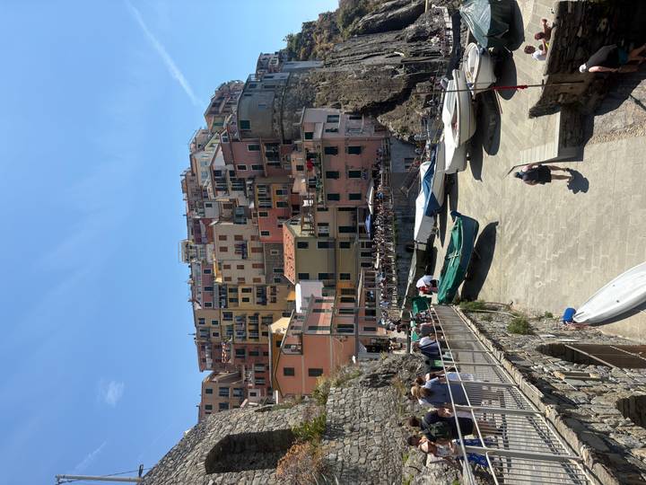 Colorful cliff-side village with small harbor, boats pulled onto a stone ramp and tourists walking along the waterfront under a bright blue sky.