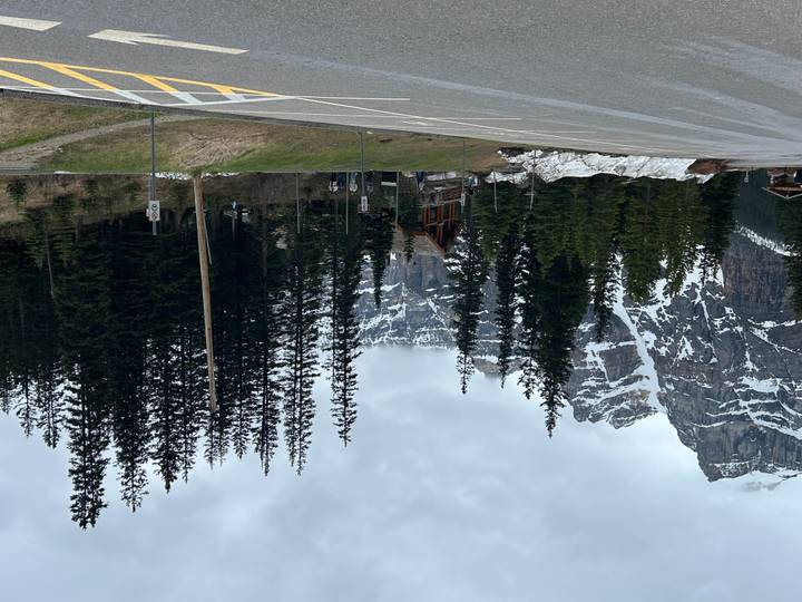 Mountain lodge nestled among tall pines with towering snow-streaked cliffs behind, viewed from a roadside parking area.