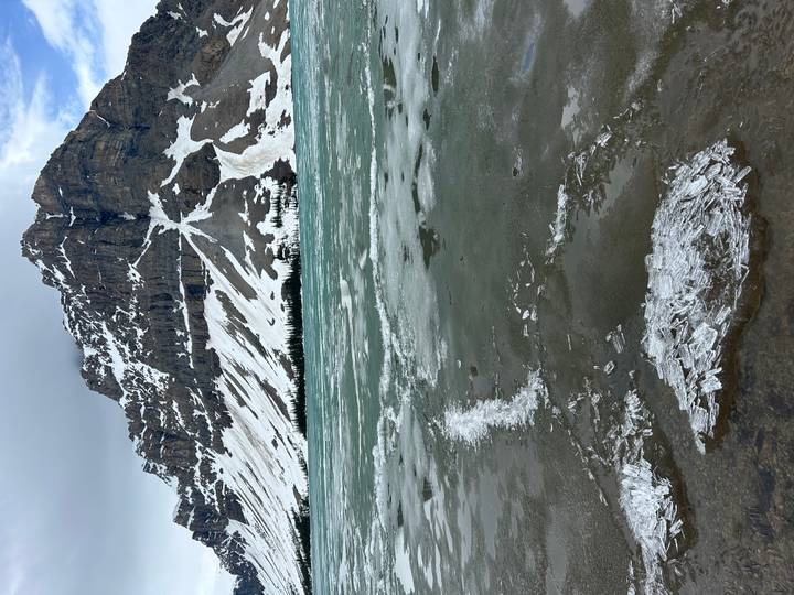 Emerald Bow Lake partially frozen with crystal ice shards in the foreground and rugged snow-striped peaks beyond.