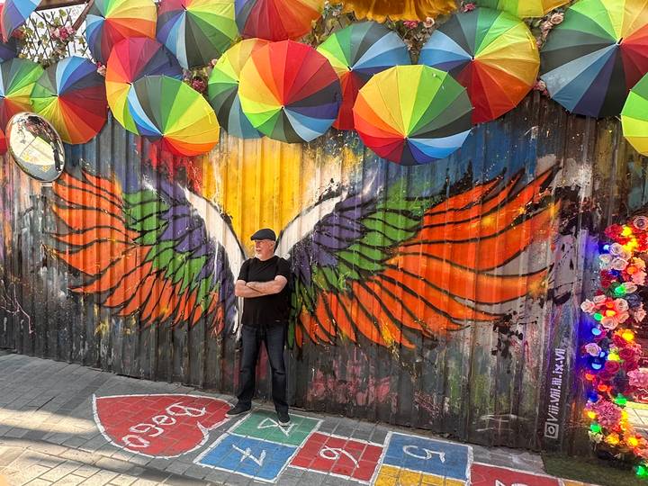 Man standing with arms crossed in front of a vibrant wing mural beneath hanging umbrellas.