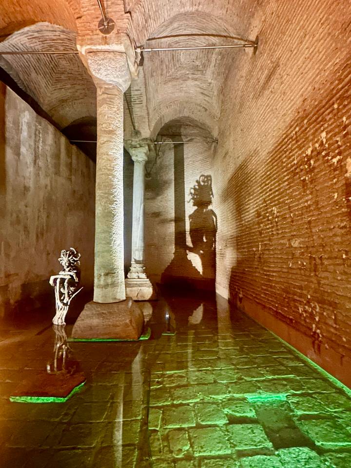 Moody interior of an ancient cistern showing columns and brick walls lit to reveal a Medusa head shadow.