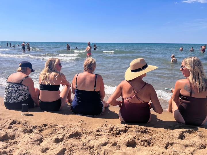 Group of women sitting on sandy shoreline chatting while beachgoers enjoy the sea behind them
