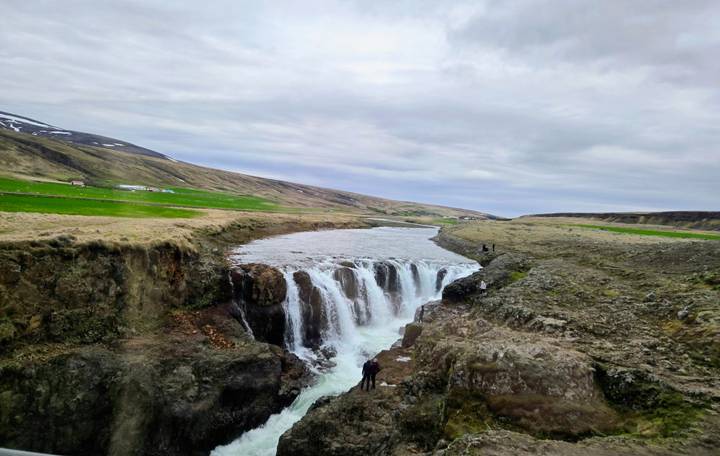 Small but powerful waterfall plunging into a rocky gorge amid Icelandic highlands with two distant visitors for scale.