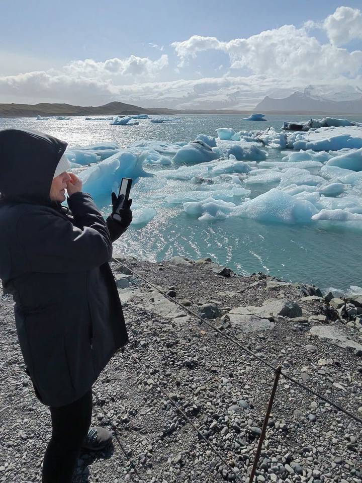 Traveller in winter coat photographing sparkling blue icebergs floating in a glacier lagoon.
