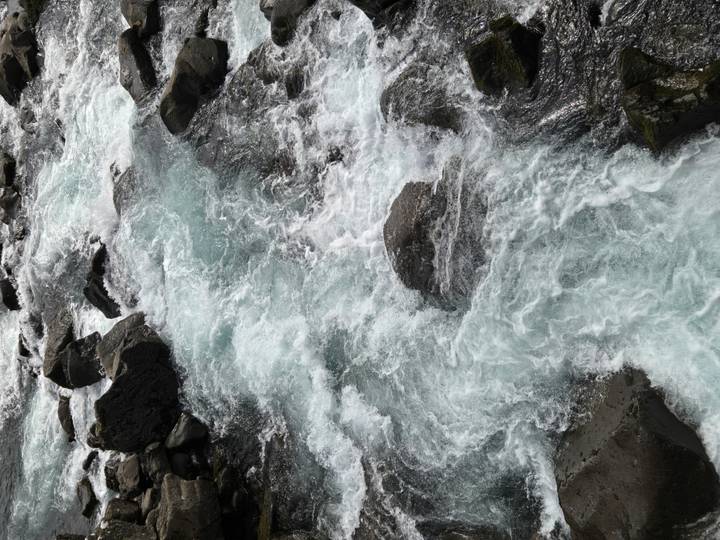Fast-flowing, foamy turquoise river water rushing over dark volcanic rocks.