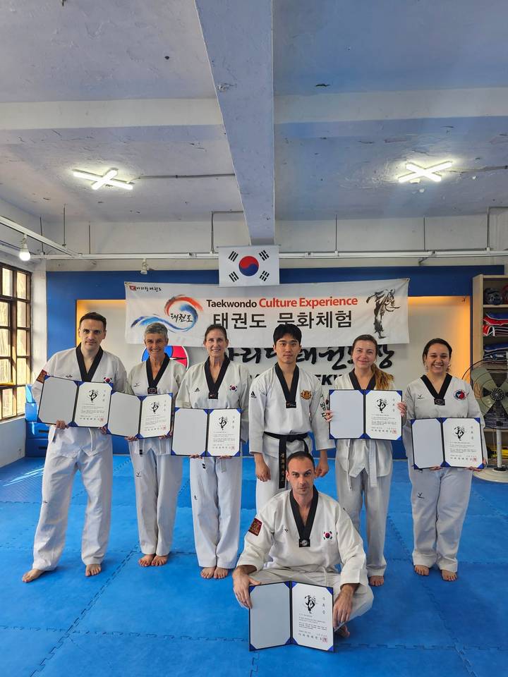 A class of taekwondo participants in uniform proudly hold certificates under a Korean flag banner inside a studio.