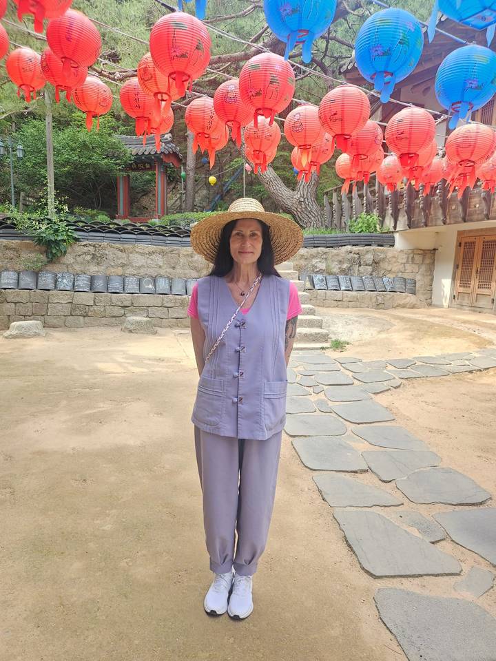A woman in a broad straw hat and simple temple-stay attire stands in a tranquil courtyard.