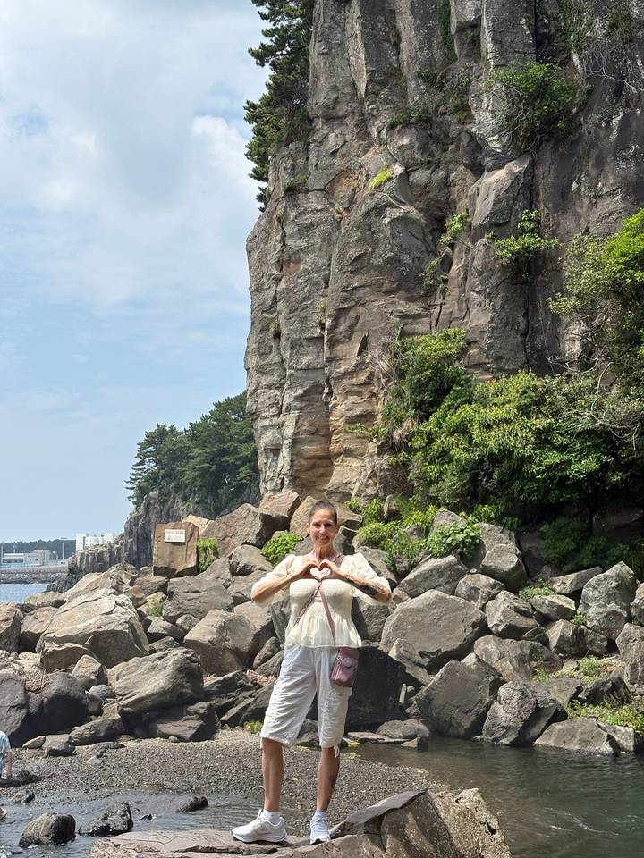 A woman forms a heart with her hands in front of rugged seaside cliffs and boulders.