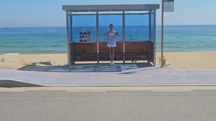 A lone traveler poses inside a glass bus stop shelter facing a wide sandy beach and blue sea.