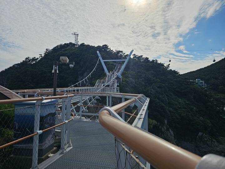 A curved pedestrian skywalk leads toward a forested hill, with cable cars visible against a blue sky.