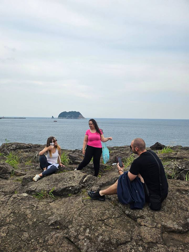 Travelers relax and take photos on rocky coastal cliffs overlooking a small offshore island.