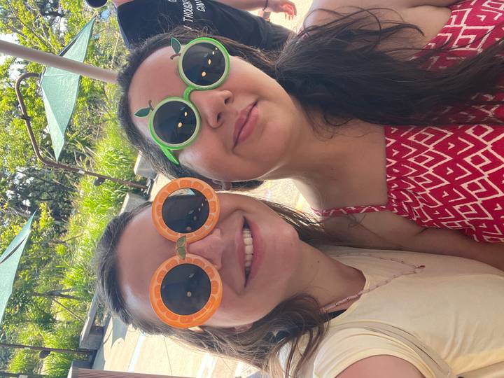 Two women smile wearing playful fruit-themed sunglasses in bright outdoor light.