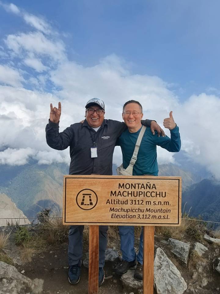 Two happy trekkers celebrate beside the wooden summit sign of Machu Picchu Mountain high above the clouds.