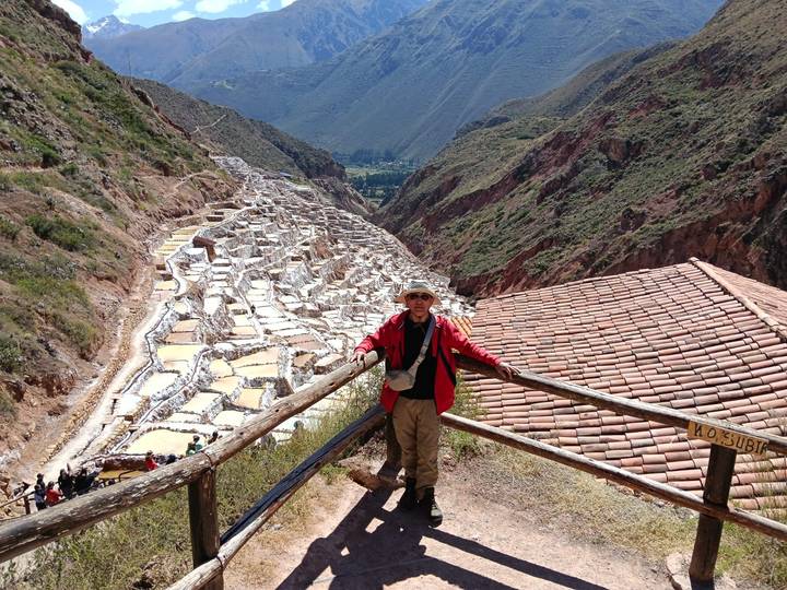 Traveler standing at a wooden railing overlooking the terraced salt ponds nestled in a steep Andean valley