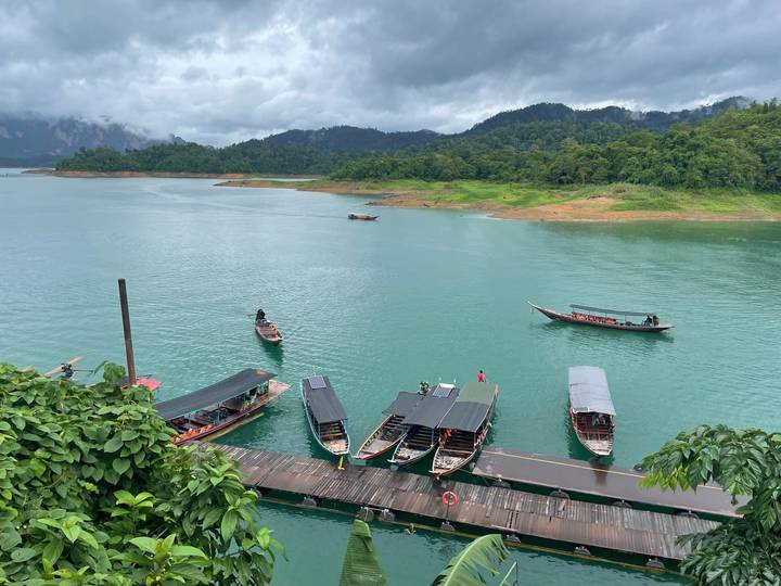 Long-tail boats moored on emerald green lake surrounded by jungle-covered limestone hills under cloudy skies.