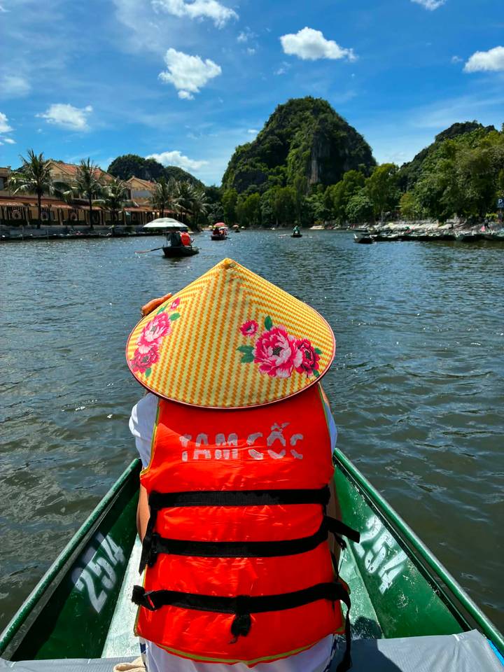 View from a rowboat of a passenger wearing a floral conical hat on a calm river.