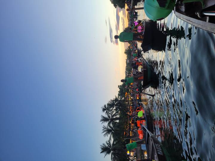 Twilight scene of rowboats on a river filled with tourists wearing life jackets amid lantern lit town.