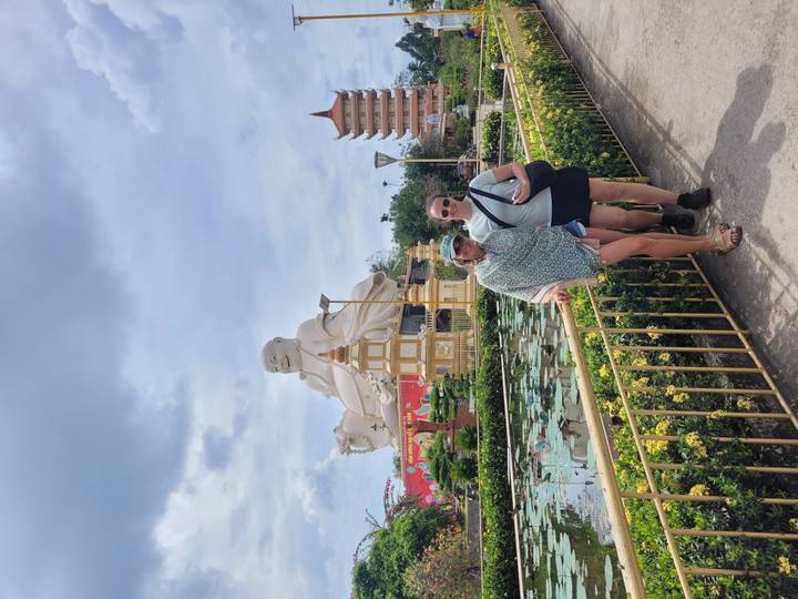 Two travellers standing before a huge seated Buddha statue and lotus pond under a cloudy sky.