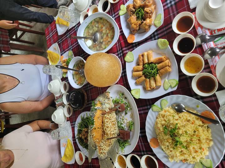 Vietnamese feast spread on a checkered table with fish, spring rolls and soups while diners sit around.