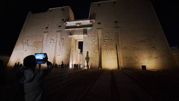 Night view of Edfu Temple illuminated with tourists capturing photos at the entrance.