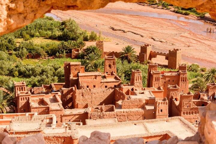 Adobe towers and walls of Ait Benhaddou rise beside a dry riverbed and palm groves.