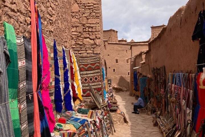 Narrow alley in Ait Benhaddou lined with vibrant Berber textiles and rugs for sale.