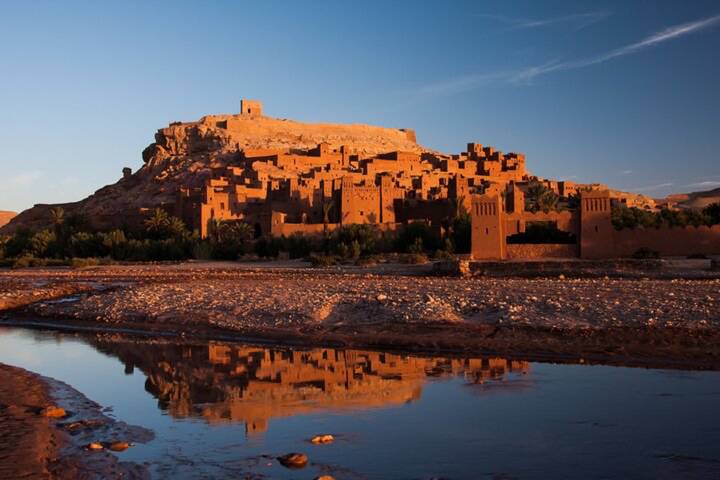 Kasbah of Ait Benhaddou reflected in the shallow river at golden sunset.