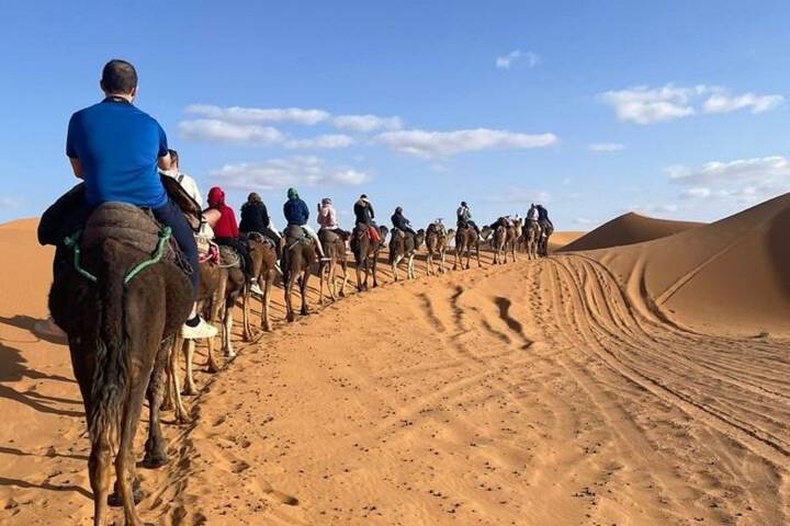 Long camel caravan follows tracks along a sandy ridge beneath scattered clouds.