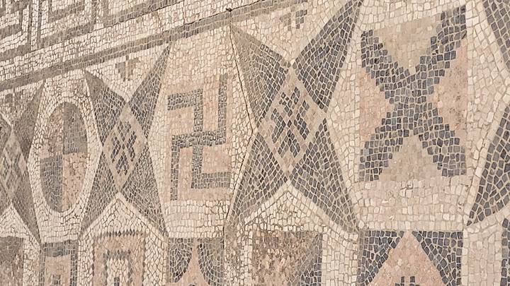 Detail of an ancient mosaic floor with geometric patterns and symbols made of small stone tiles.